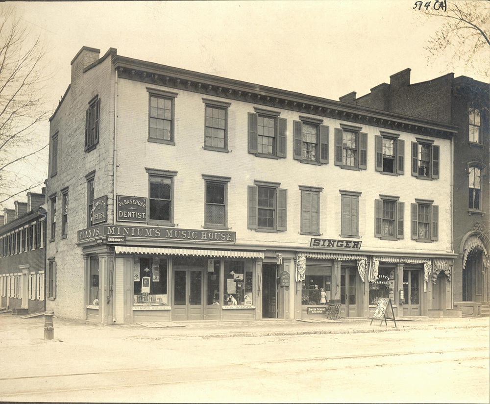 Cut stone-brick front building housing MINIUM'S MUSIC HOUSE, SINGER Sewing Machines, Dr. BASEHOAR Dentist, J. E. SIPE'S Pharmacy.